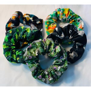 May include: A collection of five fabric scrunchies in various patterns. The scrunchies feature designs with turtles, pandas, and other animals, along with floral and abstract prints. The colors include green, black, yellow, and white.