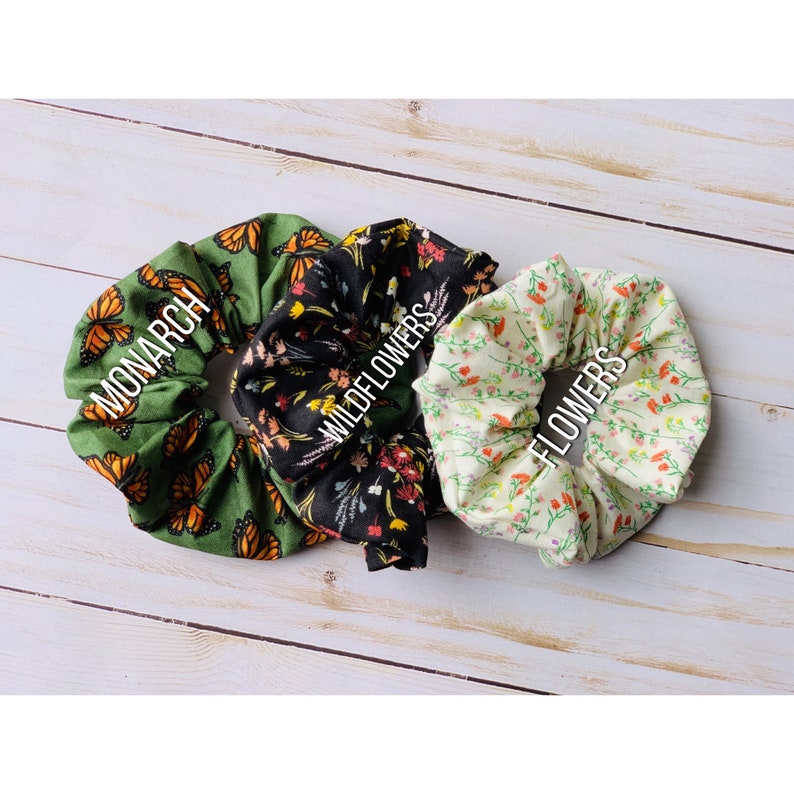 Op de afbeelding: Drie stoffen scrunchies met verschillende patronen. De eerste scrunchie is groen met een monarchvlinderpatroon en de tekst "MONARCH". De tweede scrunchie is zwart met een wilde bloemenpatroon en de tekst "WILDFLOWERS". De derde scrunchie is cr&egrave;mekleurig met een klein bloemenpatroon en de tekst "FLOWERS".