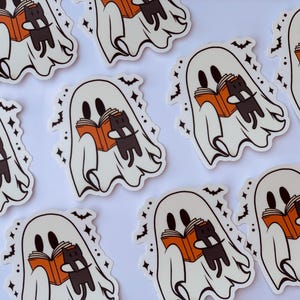 May include: A set of nine white ghost stickers with black cat and orange book illustrations. Each ghost is holding a book and a black cat is perched on top of the book. The stickers have a sparkly background.