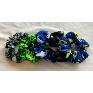 May include: Five colorful hair scrunchies with different space-themed patterns. The scrunchies are made of fabric and have a gathered, ruffled look. The patterns include aliens, stars, planets, and a black and white galaxy design.