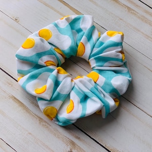 May include: A fabric scrunchie with a repeating pattern of yellow egg yolks on a white background, accented by light blue stripes and polka dots. The hair accessory is designed to gather and hold hair.