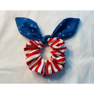 May include: A patriotic scrunchie with a bow, featuring red and white stripes and a blue section with white stars. This hair accessory is perfect for adding a touch of Americana to any hairstyle.