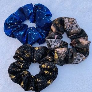 May include: Three scrunchies with celestial and star patterns. One scrunchie is navy blue with gold stars and constellations. Another scrunchie is black with gold moons and suns. The third scrunchie is black with gold floral and moon patterns.