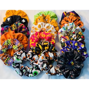 May include: A collection of Halloween-themed scrunchies in various colors and patterns. The scrunchies feature designs like pumpkins, bats, eyeballs, and floral prints. Colors include orange, black, white, green, and purple. These hair accessories are perfect for adding a festive touch.