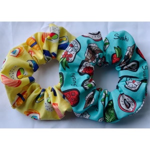 May include: Two fabric hair scrunchies with a sushi theme. One is yellow with red and black sushi rolls and the word "sushi" printed on it. The other is blue with white and red sushi rolls and the word "Sushi" printed on it.