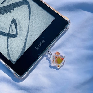 May include: A black Kindle e-reader with a clear case and a frog-themed charm attached to the bottom. The charm features a frog wearing a witch's hat and holding a wand.