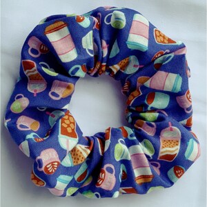 May include: A blue scrunchie with a repeating pattern of coffee cups and to-go cups in shades of pink, brown, and white. The fabric is gathered and elasticized, creating a textured, circular shape.