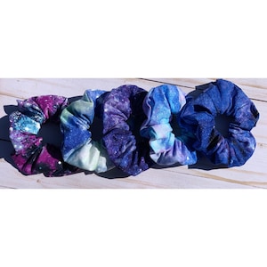 May include: Five galaxy print scrunchies in shades of blue, purple, and white. The scrunchies are made of soft fabric and are perfect for holding hair in place.