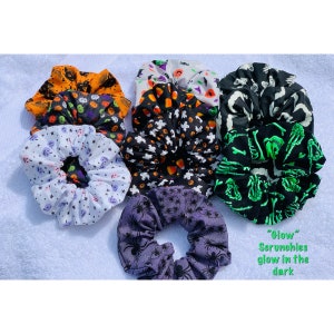 May include: A set of eight Halloween-themed scrunchies in various colors and patterns, including black, white, orange, green, and purple. The scrunchies feature designs such as ghosts, pumpkins, candy corn, spiders, and skeletons. The text "Glow" Scrunchies glow in the dark" is written below the scrunchies.
