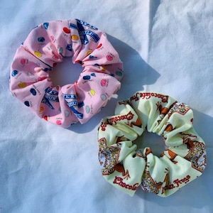 May include: Two fabric scrunchies. One is pink with a pattern of jelly beans and text. The other is cream-coloured with a chocolate frog design and the text "Chocolate Frogs". These hair accessories are perfect for adding a touch of fun to any hairstyle.