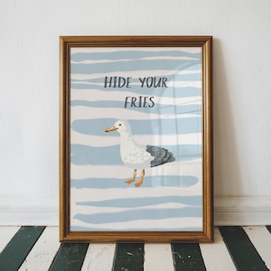 May include: A framed print featuring a cartoon seagull with the text "HIDE YOUR FRIES" above it. The background has blue and white horizontal stripes. The seagull is white with gray and black accents and orange feet and beak. The frame is brown.