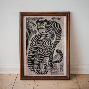 May include: A framed black and white print of a tiger with intricate details. The tiger is sitting with its legs crossed and its tail curled around its body. The print is on a light gray background and has a brown wooden frame.