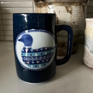 May include: A navy blue ceramic mug with a white oval design featuring a blue and white duck illustration. The duck has a patterned body with hearts and anchors. The mug has a dark blue handle.