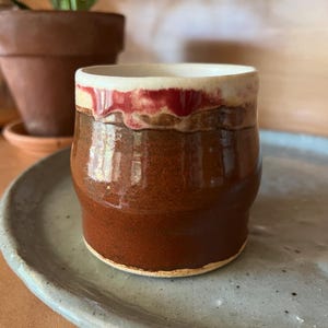 Könnte beinhalten: Handgefertigte Keramiktasse mit glänzend brauner Glasur. Der Rand hat ein rotes und weißes Band, das einen Kontrast bildet. Die Tasse steht auf einem hellblauen, gesprenkelten Keramikteller, was eine rustikale, handwerkliche Ästhetik suggeriert.