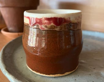 Könnte beinhalten: Handgefertigte Keramiktasse mit glänzend brauner Glasur. Der Rand hat ein rotes und weißes Band, das einen Kontrast bildet. Die Tasse steht auf einem hellblauen, gesprenkelten Keramikteller, was eine rustikale, handwerkliche Ästhetik suggeriert.