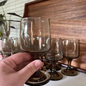 May include: A collection of brown-tinted wine glasses. The glasses have a rounded bowl and a stem. The glasses are arranged on a white surface with a wooden tray in the background. The glasses are made of glass.