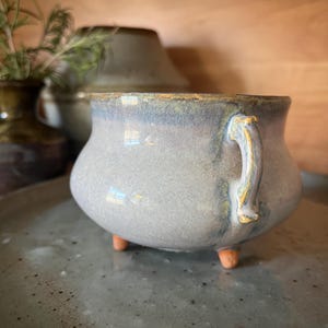 May include: A small, round, ceramic pot with a light blue glaze and a single handle. The pot has three small, brown feet. The rim has a gold-colored accent. The pot is sitting on a gray, speckled surface.