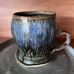 May include: A handmade ceramic mug with a blue and brown glaze. The mug has a rounded shape, a small handle, and sits on a matching saucer. The glaze has a dripped effect, creating a unique texture. The mug is suitable for coffee or tea.