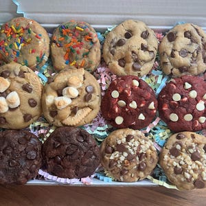 May include: A selection of a dozen cookies in a pink box with colourful paper. The cookies include chocolate chip, red velvet, and sprinkle-covered varieties. Some cookies have marshmallows, nuts, and chocolate chunks.