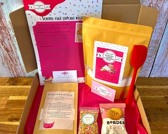 Puede incluir: Una caja rosa y blanca llena de ingredientes para hacer cupcakes. La caja incluye una bolsa de mezcla para pastel, una bolsa de chispas, una bolsa de remolinos de galletas, una espátula y una tarjeta de receta. La caja está etiquetada como "School Cake Cupcake Mix".