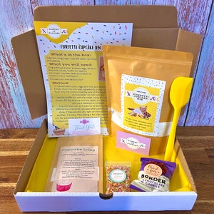 May include: A yellow box containing a funfetti cupcake baking kit. The kit includes a bag of cake mix, a bag of sprinkles, a spatula, a bag of chocolate chips, and a recipe card. The box is labelled "Funfetti Cupcake Recipe".