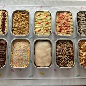 May include: Ten rectangular foil containers filled with different frosted cakes. The cakes have varied toppings, such as red velvet, caramel swirls, carrot cake with carrot decorations, and chocolate with sprinkles. The cakes are on a white surface.