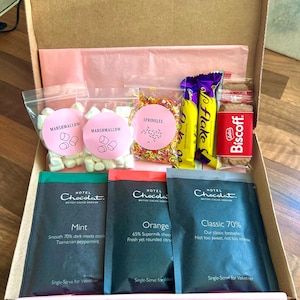 Pode incluir: Uma caixa de cartão cheia de um conjunto de presente de chocolate quente. O conjunto inclui duas sacolas de marshmallows, uma sacola de granulado, duas barras de chocolate, um pacote de bolachas Biscoff e três pacotes individuais de chocolate quente nos sabores: Menta, Laranja e Clássico 70%.