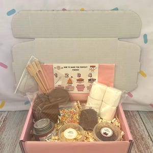 Może przedstawiać: Różowe pudełko wypełnione składnikami na s'mores, w tym krakersami Grahama, piankami marshmallow, czekoladą, posypką i małą puszką. Karta głosi: "Jak zrobić idealne s'mores". Dołączone są drewniane szpikulce.