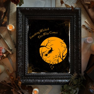 May include: A rectangular black ornate frame containing a print with a large orange moon, a silhouette of a witch flying on a broomstick, and bare tree branches. The text "Something Wicked This Way Comes" is printed in gold in the upper left corner.