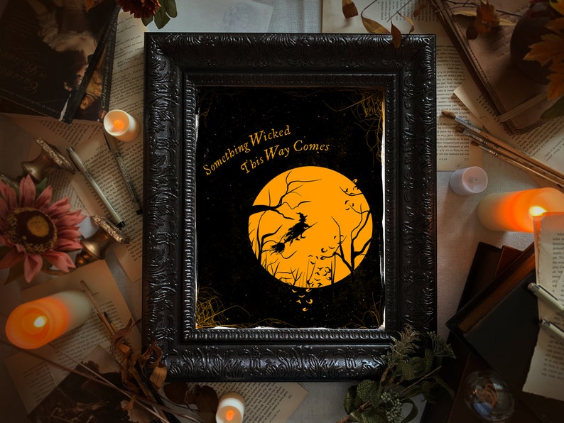May include: A rectangular black ornate frame containing a print with a large orange moon, a silhouette of a witch flying on a broomstick, and bare tree branches. The text "Something Wicked This Way Comes" is printed in gold in the upper left corner.
