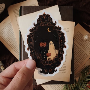May include: A die-cut sticker featuring a black ornate frame containing a white ghost holding a small orange jack-o'-lantern, a larger jack-o'-lantern, and a crescent moon against a dark background with subtle stars, all enclosed in a white border.