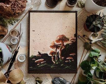 Könnte beinhalten: Gerahmtes Kunstwerk mit braunen und beigen Pilzen mit grünem Moos auf beigem Hintergrund, in einem dunkelbraunen Rahmen. Das Kunstwerk ist von Pflanzen, Schreibwaren und dekorativen Gegenständen umgeben.