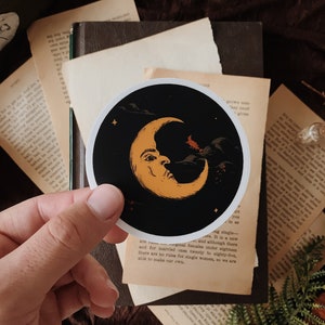 May include: A circular sticker featuring a yellow crescent moon with a human-like face set against a black night sky. The design includes a small red silhouette of a witch flying on a broomstick, dark clouds, and tiny yellow stars scattered across the background.