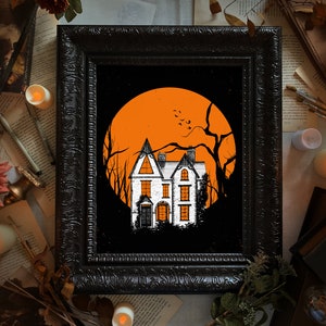 May include: A framed art print featuring a white house with orange-lit windows set against a large, solid orange circular moon. The scene includes black silhouettes of bare, leafless trees and a dark, textured frame surrounding the illustration.