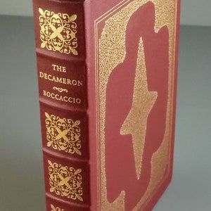 May include: A burgundy hardcover book titled "The Decameron Boccaccio" with gold gilded detailing. The spine features ornate gold designs. A gold bookmark is visible. The book's cover has a gold design.