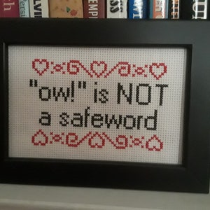 May include: A framed cross-stitch with a white background and black text that reads "'ow!' is NOT a safeword". The text is surrounded by a border of red hearts.