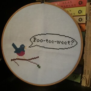 May include: A white embroidery hoop with a blue bird on a branch, saying "Poo-tee-weet?" in black thread.