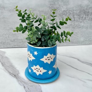 May include: A cylindrical ceramic planter with a bright blue glaze featuring white cloud shapes with smiling faces and small white stars. The planter sits on a matching blue circular saucer and contains a green leafy plant with small round leaves.