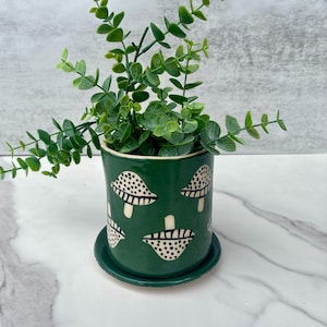 May include: A cylindrical green ceramic planter with a matching saucer, featuring a repeating pattern of white mushrooms with black polka dots. A vibrant green artificial plant with small, rounded leaves is potted inside the container.