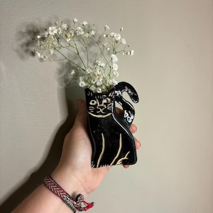 May include: A black ceramic cat-shaped vase with white accents, holding white flowers. The vase has a detailed cat face and a curved tail. The vase is approximately 6 inches tall.
