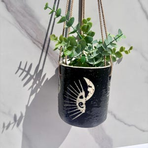 May include: A cylindrical black ceramic hanging planter with a speckled finish and a white sun and moon graphic design. The planter is suspended by thin tan cords and contains a green leafy plant with small rounded leaves extending upwards.