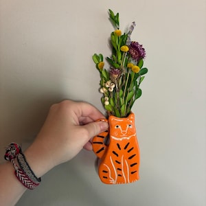 May include: Orange ceramic cat-shaped vase with black stripes and white details, holding a bouquet of dried flowers. The vase has a handle and is held by a hand. The flowers include yellow, purple, and green elements.