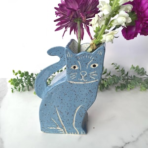 May include: A blue ceramic cat-shaped vase with a speckled finish, holding purple and white flowers. The vase has a whimsical design with carved facial features and a curved tail. The flowers add a pop of color against the blue.