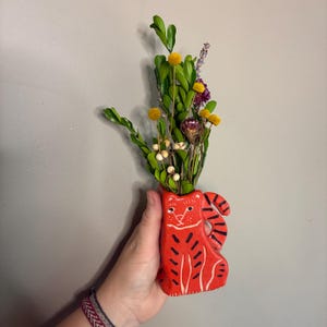 May include: A small, red ceramic vase in the shape of a tiger, holding a bouquet of dried flowers and greenery. The vase has black stripes and white details. The bouquet includes yellow, purple, and white flowers.