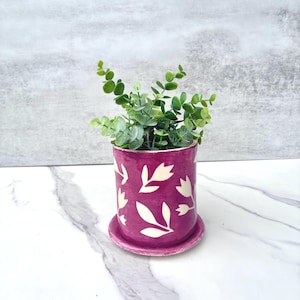 Pode incluir: Um vaso cilíndrico de cerâmica num tom magenta vibrante com um padrão floral branco pintado à mão. O vaso assenta num pires circular a condizer e contém uma pequena planta verde artificial com folhas arredondadas.