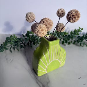 Puede incluir: Un pequeño jarrón de cerámica verde lima con un diseño de sol, que contiene varios tallos de flores secas de color beige. El jarrón tiene una parte superior cuadrada y una superficie texturizada. La vegetación decorativa está en el fondo.