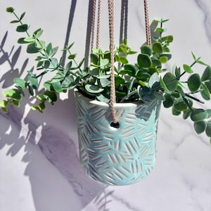 May include: A light blue ceramic hanging planter with a textured dash pattern, suspended by thin beige cords. The planter contains a green artificial eucalyptus plant with multiple stems and rounded leaves, creating a full, trailing appearance.