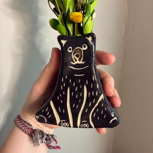 May include: Black ceramic bear-shaped vase with a whimsical design. The vase features a bear face and body with white accents. The vase is filled with green foliage and a yellow flower. A unique decorative item.