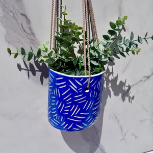 Puede incluir: Una maceta colgante de cerámica azul vibrante con un patrón de rayas blancas grabadas. El recipiente cilíndrico está suspendido por cuerdas trenzadas de color marrón claro y contiene una planta artificial verde con tallos.