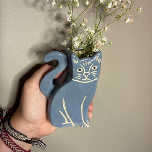May include: A blue ceramic cat-shaped vase with white detailing, filled with white flowers. The vase features a cat design with a tail and face. The flowers provide a natural contrast to the decorative item.